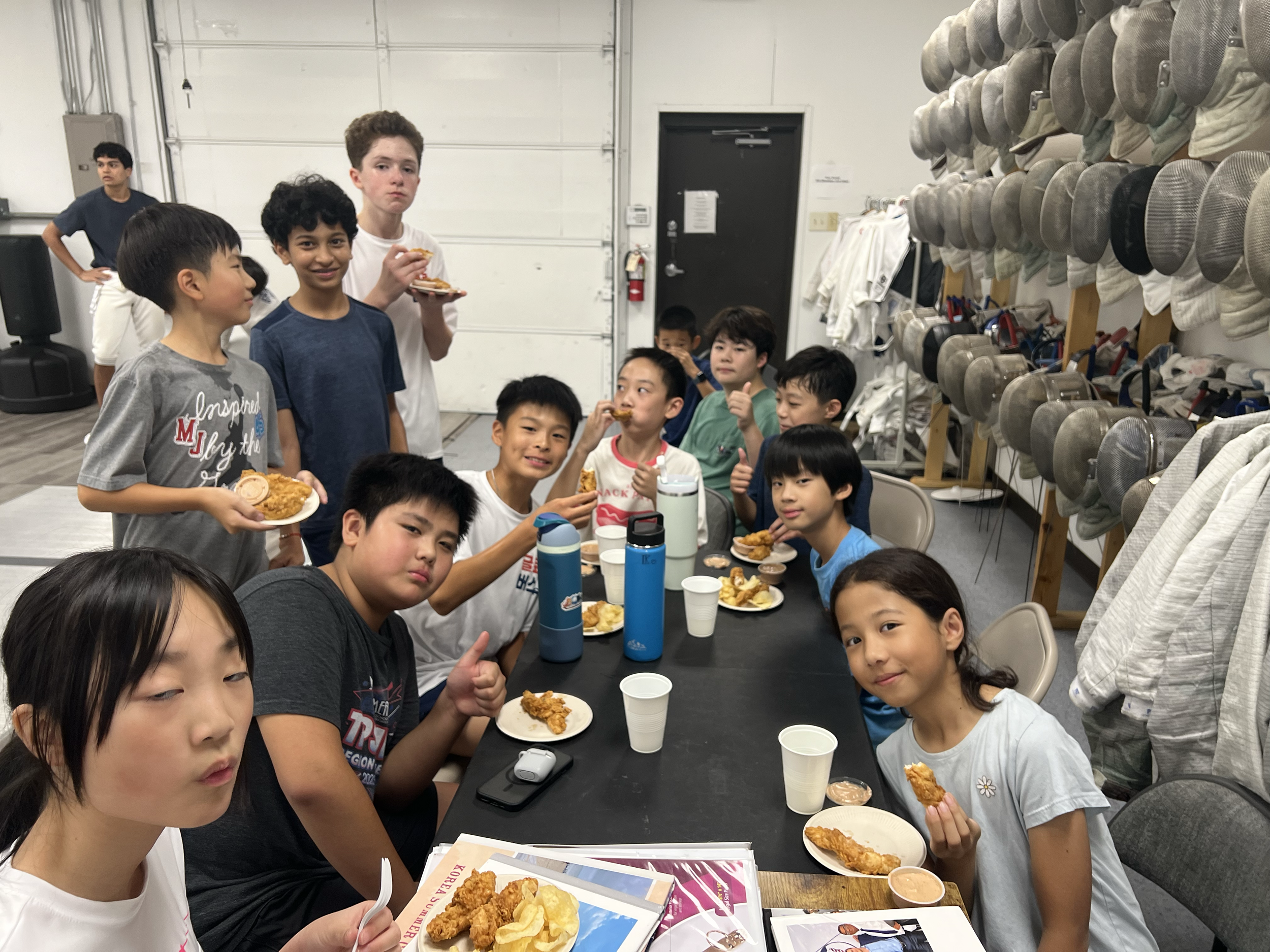 A group of children gathered around a table eating fried chicken in a room with fencing gear stored on the wall.
