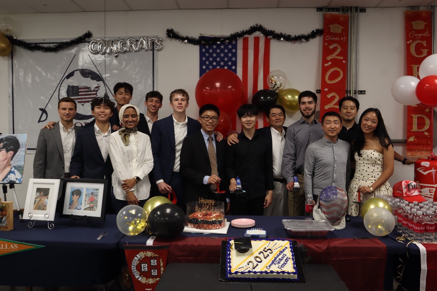 Group of thirteen people celebrating a graduation in a decorated room with banners, balloons, and flags, including an American flag and a red banner that says "Class of 2022 D." There are graduation-themed decorations, photos, and a cake with "2022" 
