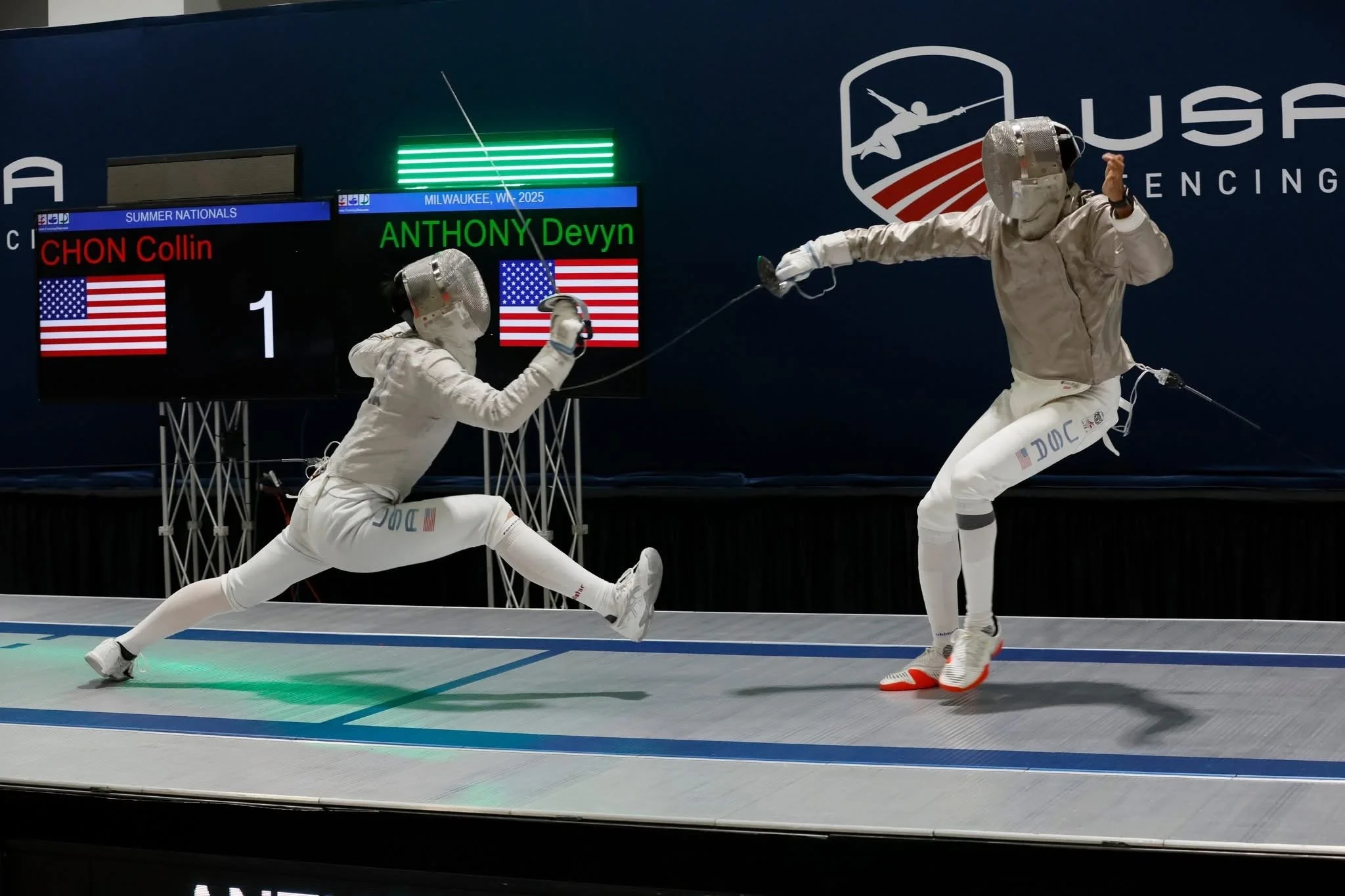 Two fencers in white protective gear engaging in a match, with one lunging towards the other on a fencing strip, with a scoreboard displaying names 'CHON Collin' and 'ANTHONY Devyn' and flags, in front of a backdrop with USA fencing logo.