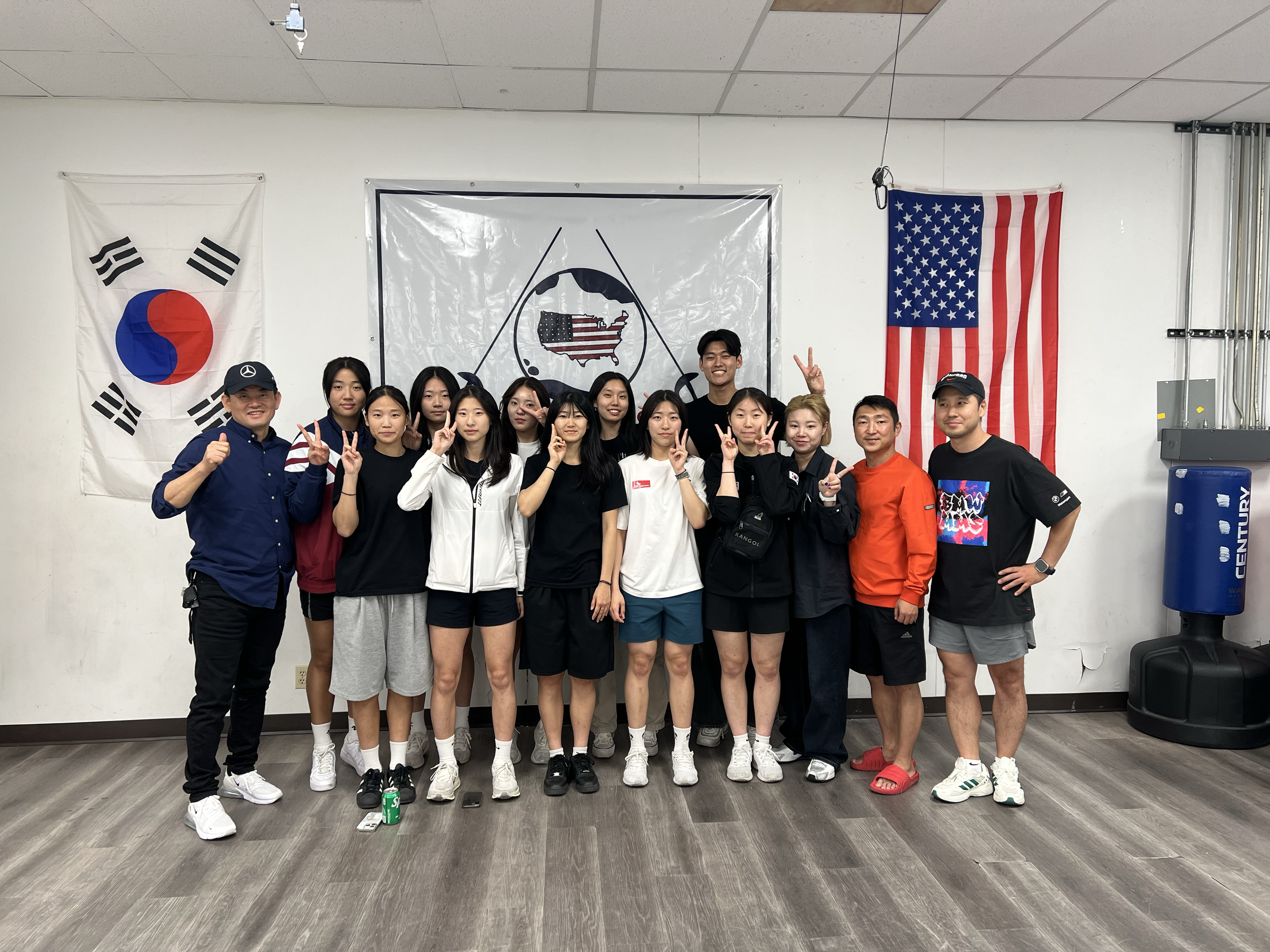 Group of young people and two adults posing for a photo in front of flags of South Korea and the United States, with a world map graphic in between, inside a room with a wooden floor and white walls.