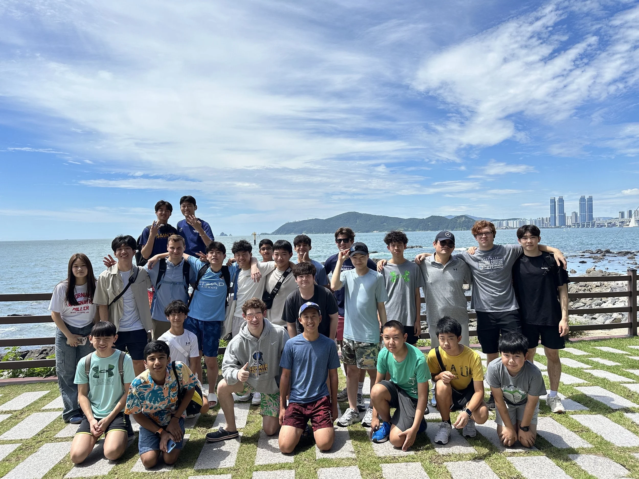 Group of young people posing outdoors near the water with city skyline and mountains in the background.
