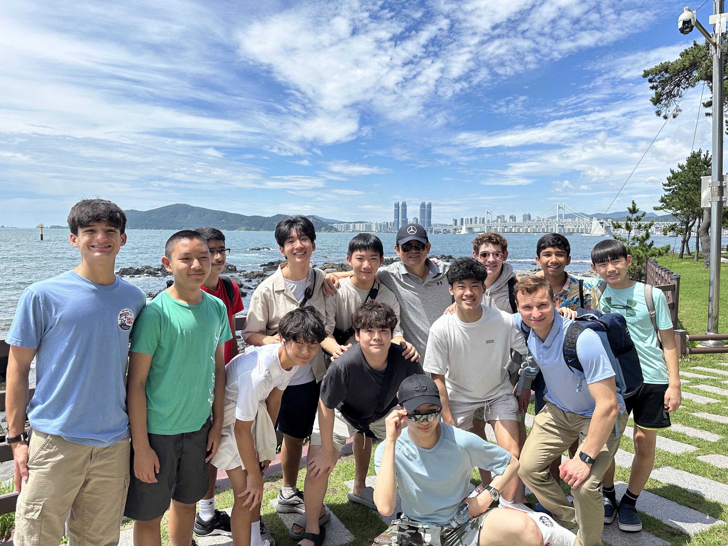 Group of teenagers and adults posing outdoors by the water with a cityscape and bridge in the background on a sunny day.