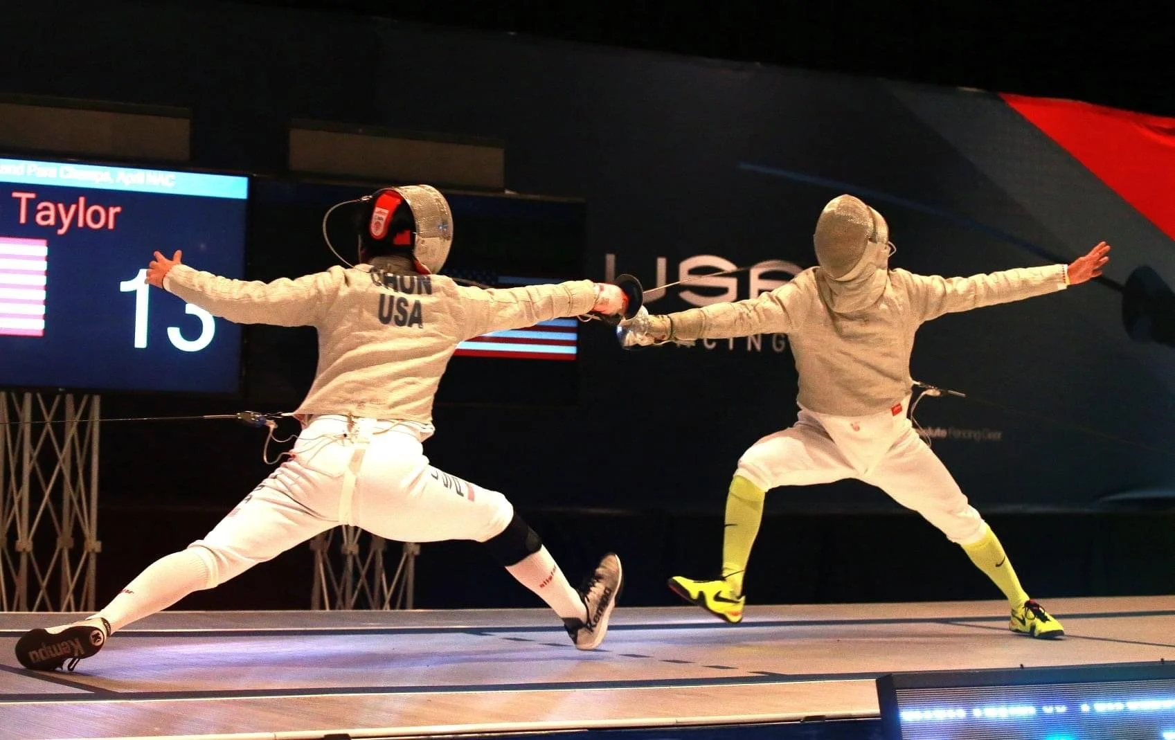 Two fencers engaged in a match, both dressed in fencing gear, with their arms extended and holding fencing swords. The fencer on the left has 'Chun USA' on their back, and there is a scoreboard in the background showing the name 'Taylor' and the scor