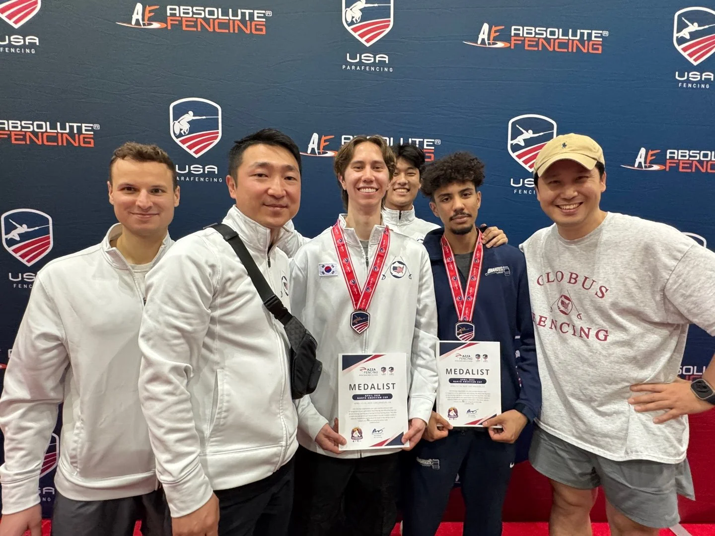 Group of five male fencers in white and dark jackets with medals and certificates standing in front of a blue backdrop with 'Absolute Fencing' and 'USA ParaFencing' logos.