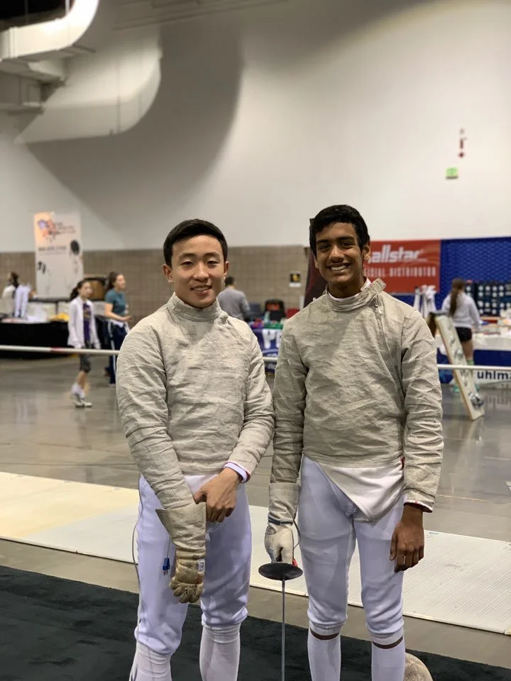 Two male fencers in fencing uniforms, smiling, standing on a fencing strip with their masks off, holding their fencing swords.