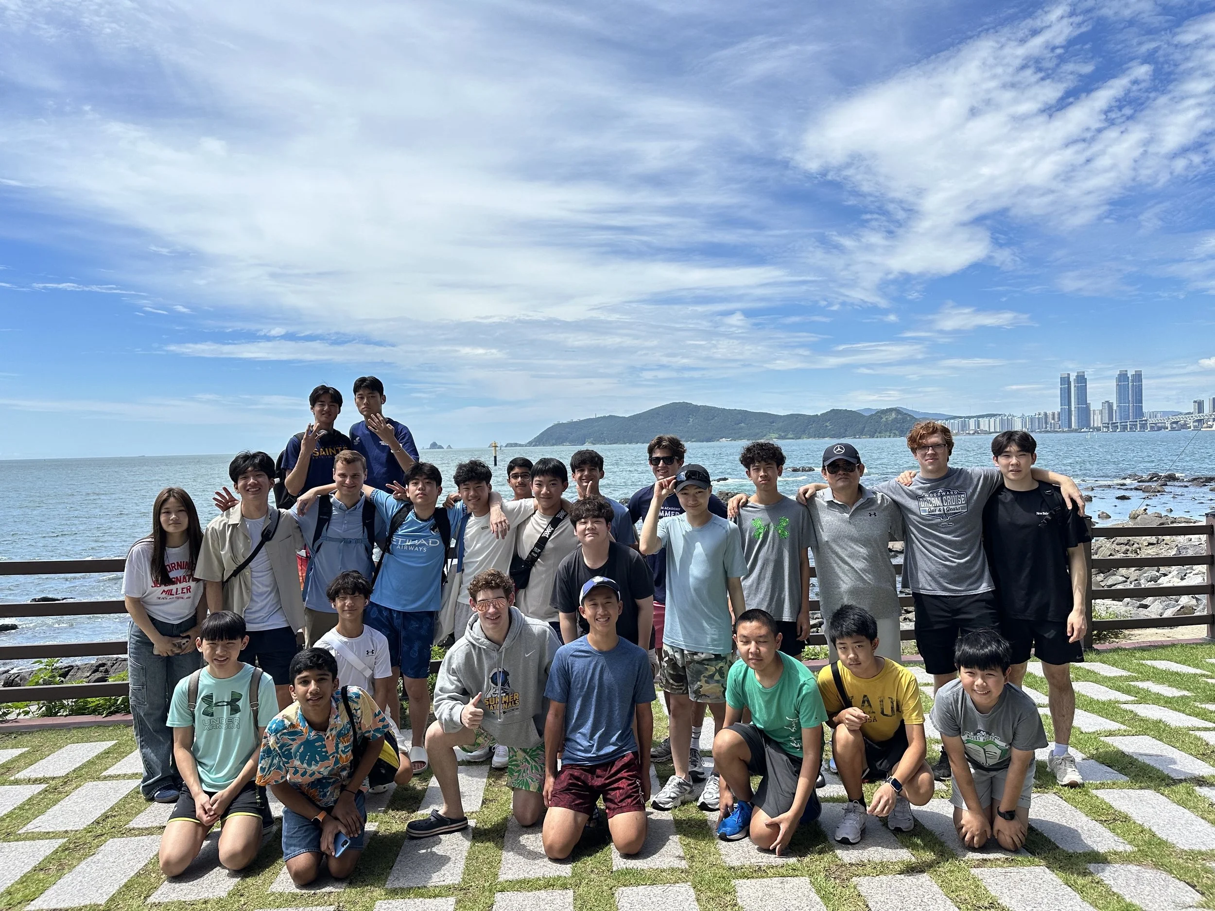 Group of young people and a woman posing outdoors near the water with a city skyline and green hills in the background.
