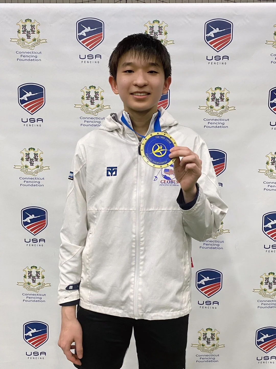 Young male athlete holding a gold medal at a fencing competition, standing in front of a backdrop with logos of USA Fencing and Connecticut Fencing Foundation.