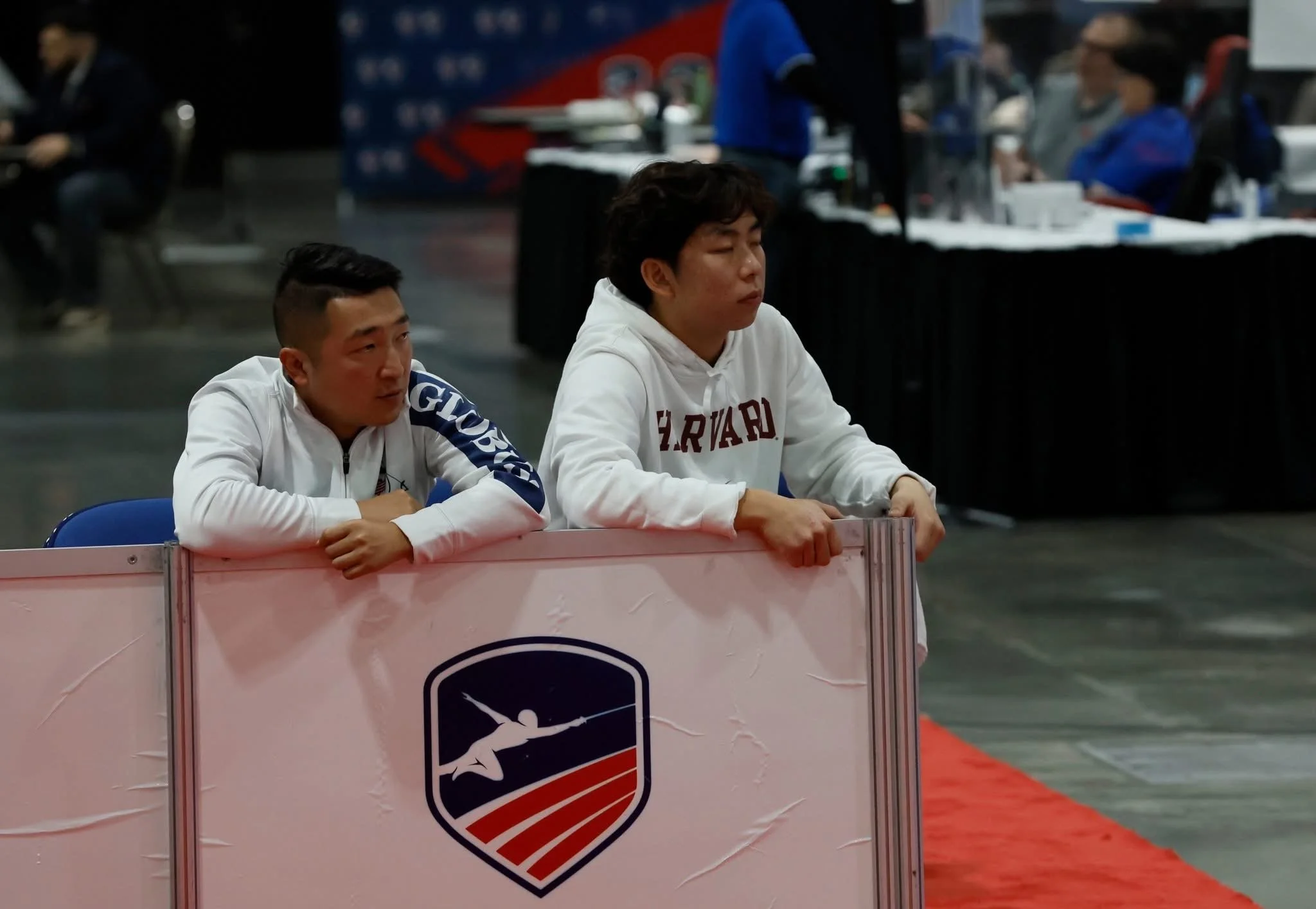 Two young men leaning on a barrier with a fencing-related logo, observing an event.