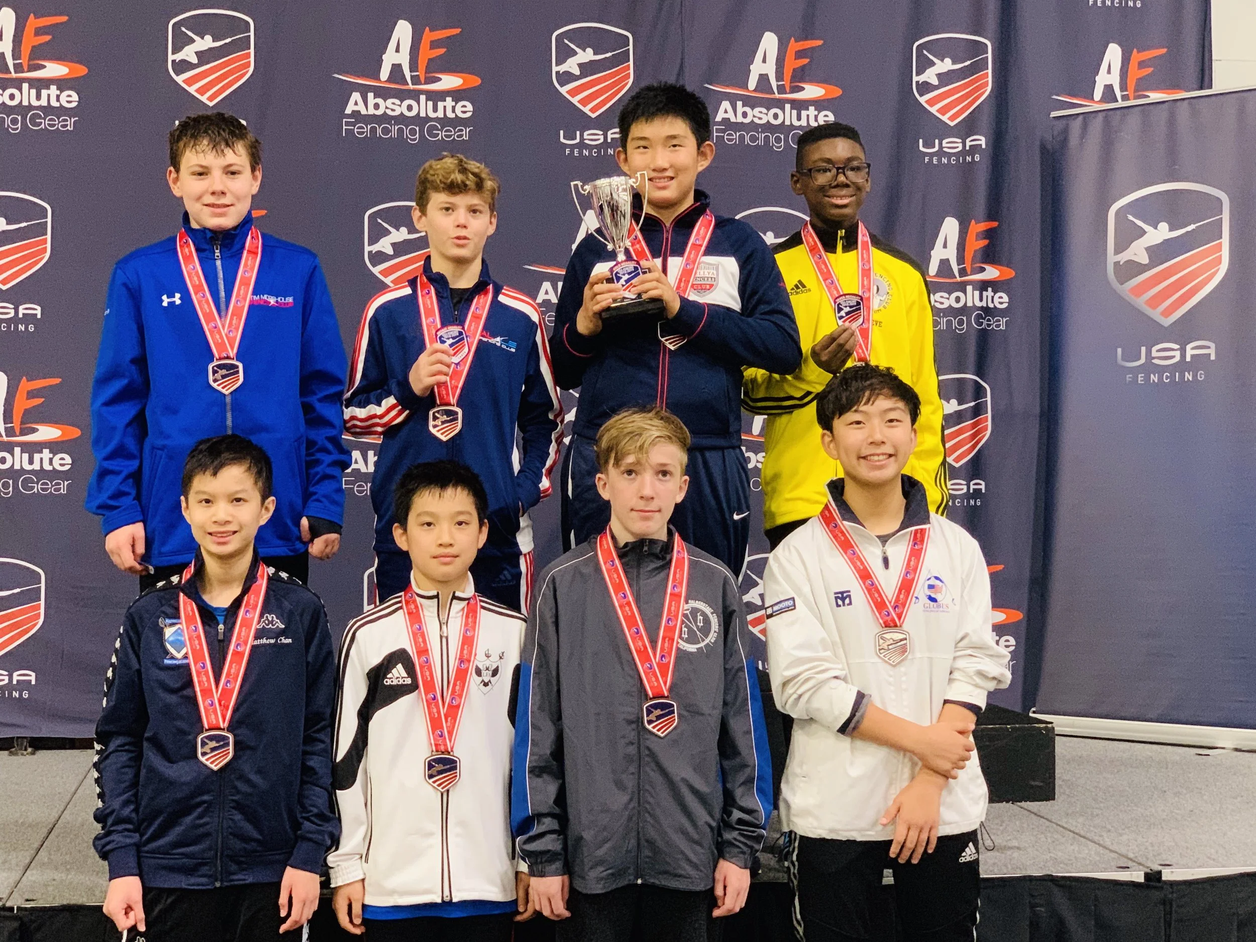 Young boys standing on a podium receiving medals at a fencing competition, with one holding a trophy, against a backdrop displaying the logos 'Absolute Fencing Gear' and 'USA Fencing'.