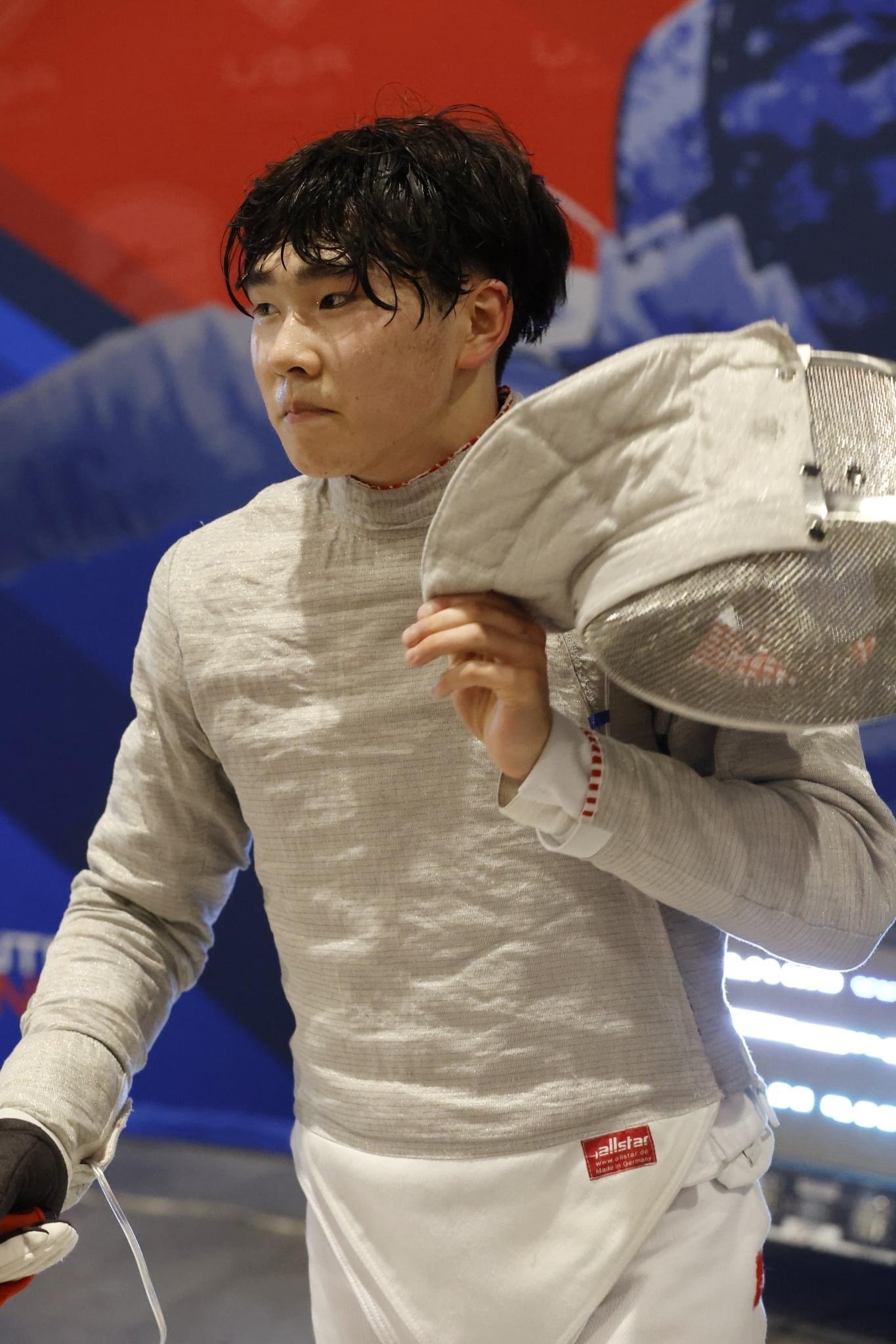 Young male fencer walking with his mask in his left hand and a small electronic device in his right hand, wearing fencing gear, in an indoor sports arena.