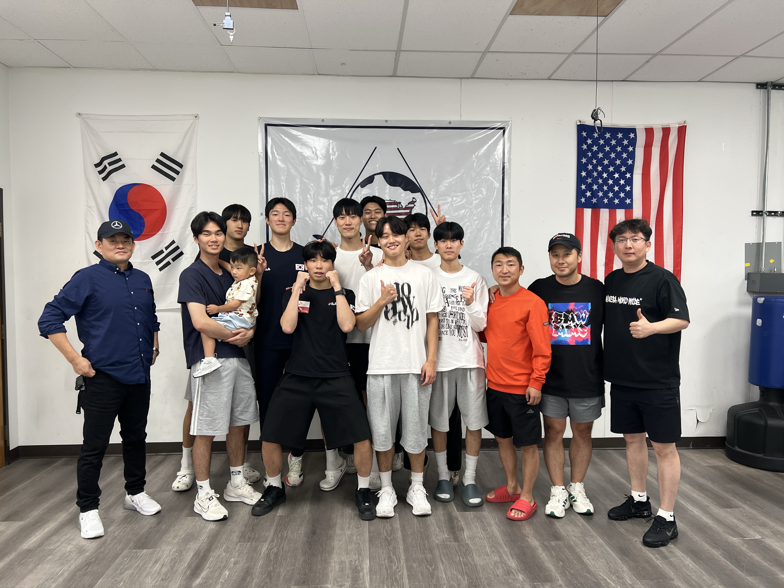 Group of people posing indoors with flags of South Korea and the United States hanging on the wall behind them. The group includes adults and children, some making peace signs.