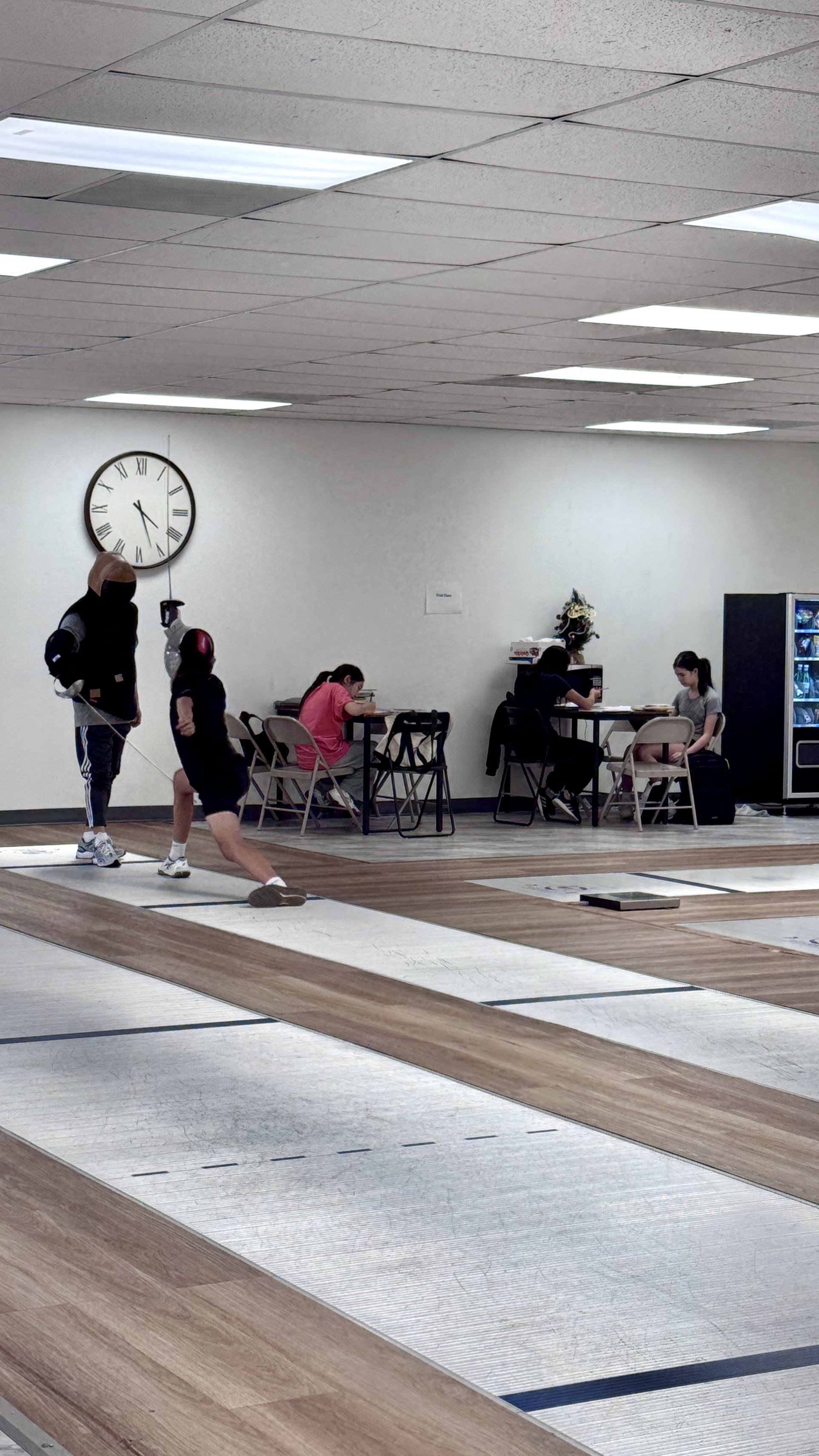 People practicing fencing in an indoor gym while two girls read at tables in the background.