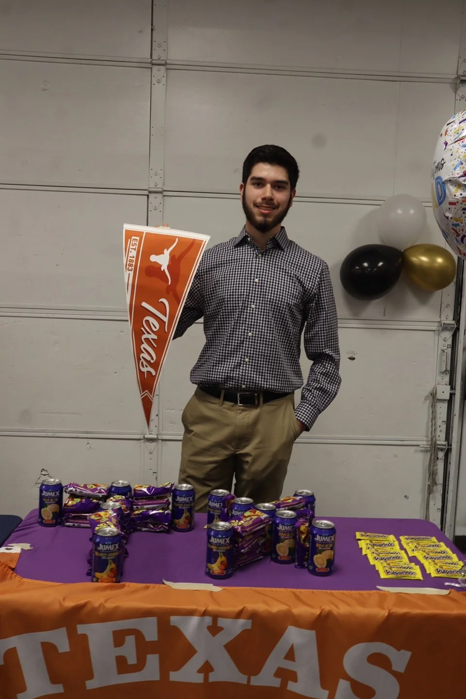 A young man standing behind a table with snacks and drinks, holding a Texas Longhorns pennant, at a celebration with balloons.
