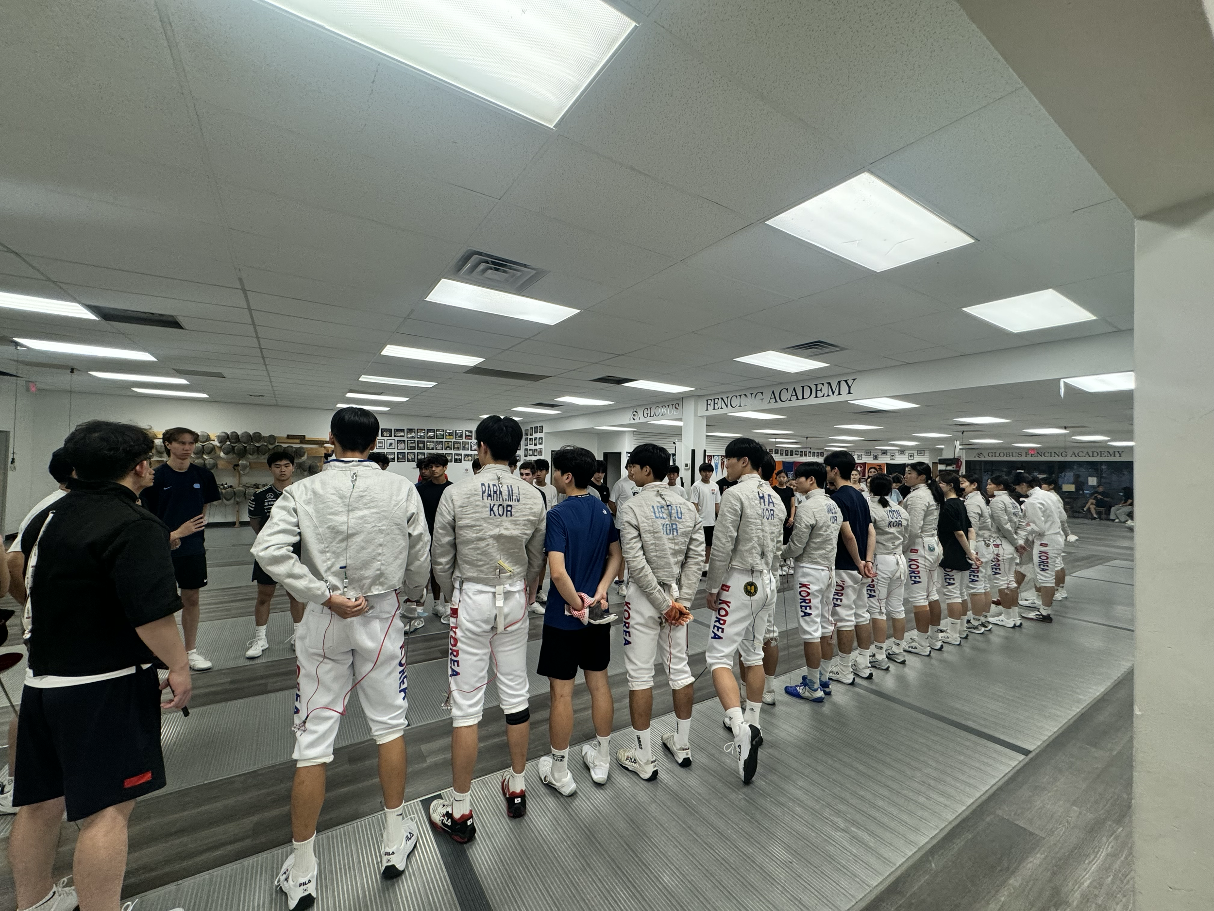 A group of fencers in white gear with 'KOREA' written on their pants stands in line inside fencing training facility, listening to instructor.