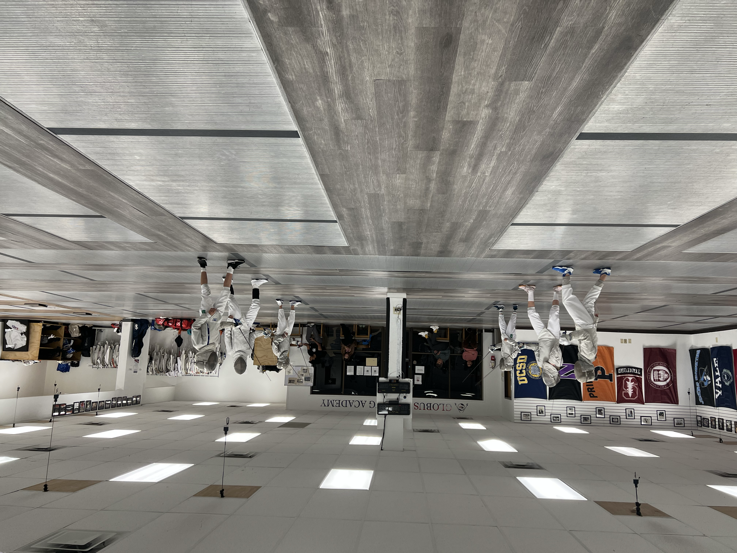 Group of people in white martial arts uniforms and protective gear practicing fencing in a sports hall, with banners hanging on the wall.
