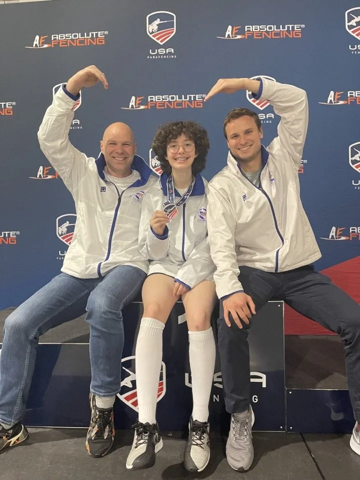 Three fencers sitting on a podium in front of a backdrop with 'Absolute Fencing' and 'USAF Parafencing' logos. They are smiling, with the person in the middle holding a medal.
