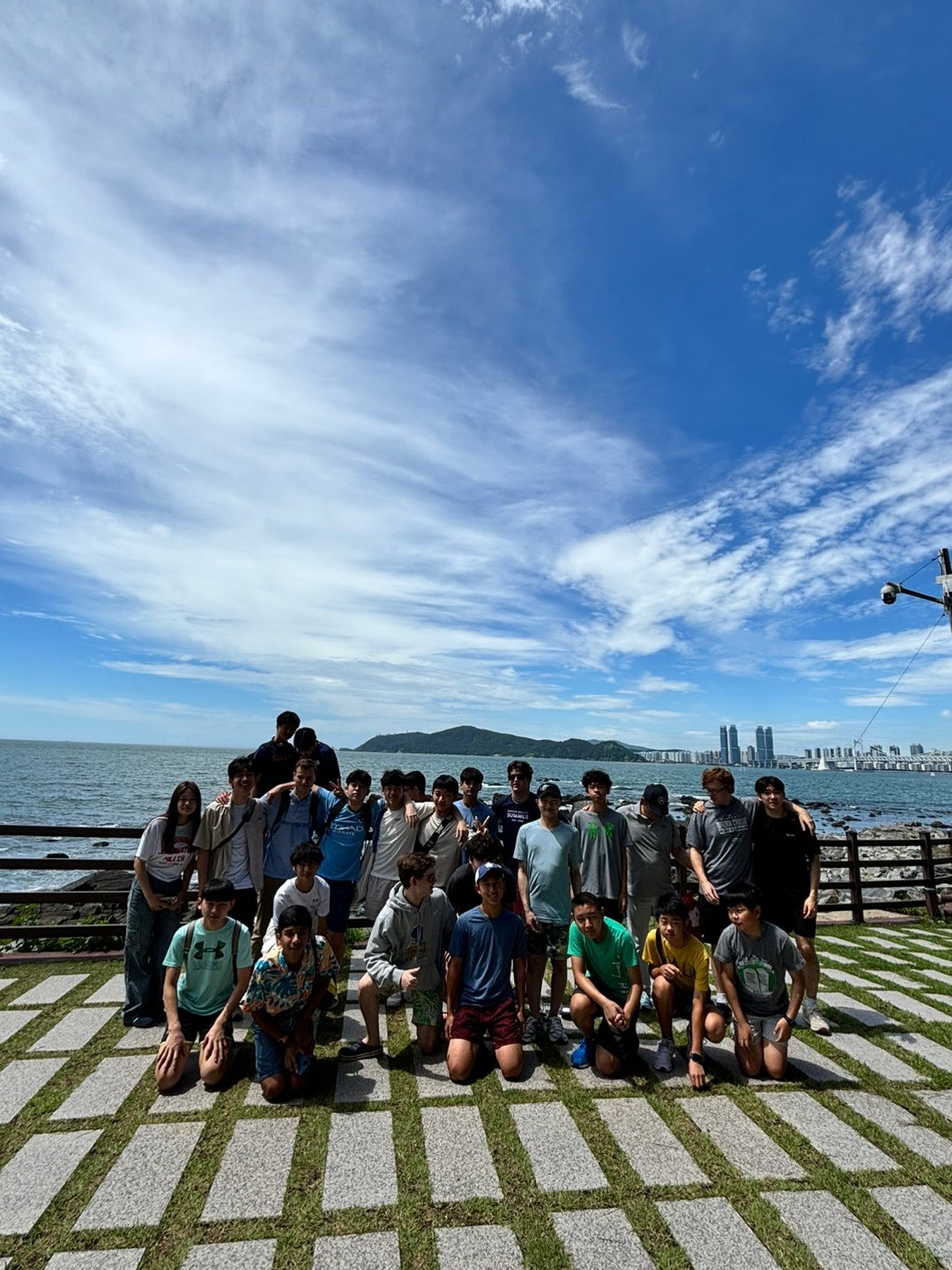Group of young people posing for a photo at the seaside with a cityscape and mountains in the background, under a partly cloudy blue sky.
