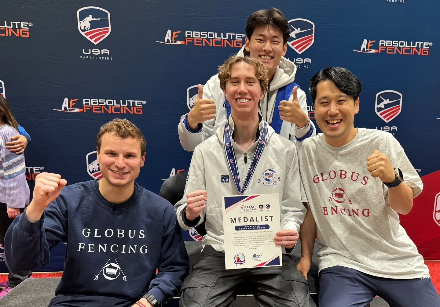 Four young men celebrate at a fencing event with medals and a certificate, in front of a backdrop with 'Absolute Fencing' and 'USA ParaFencing' logos. One holds a 'Medalist' certificate.