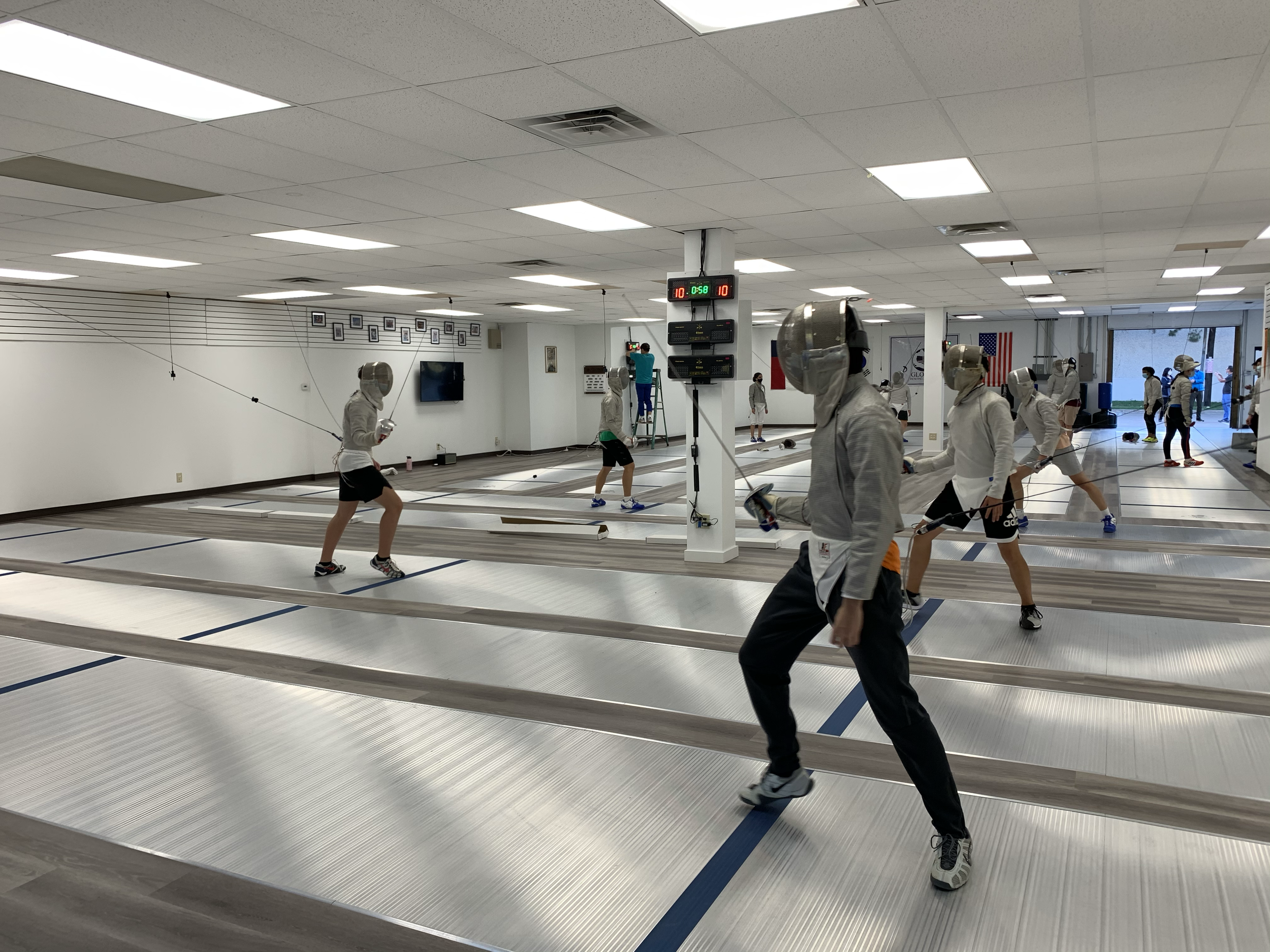 Indoor fencing facility with several fencers practicing with protective gear and masks on,  fencing strips marked on the floor, and a digital scoreboard displaying scores and time.