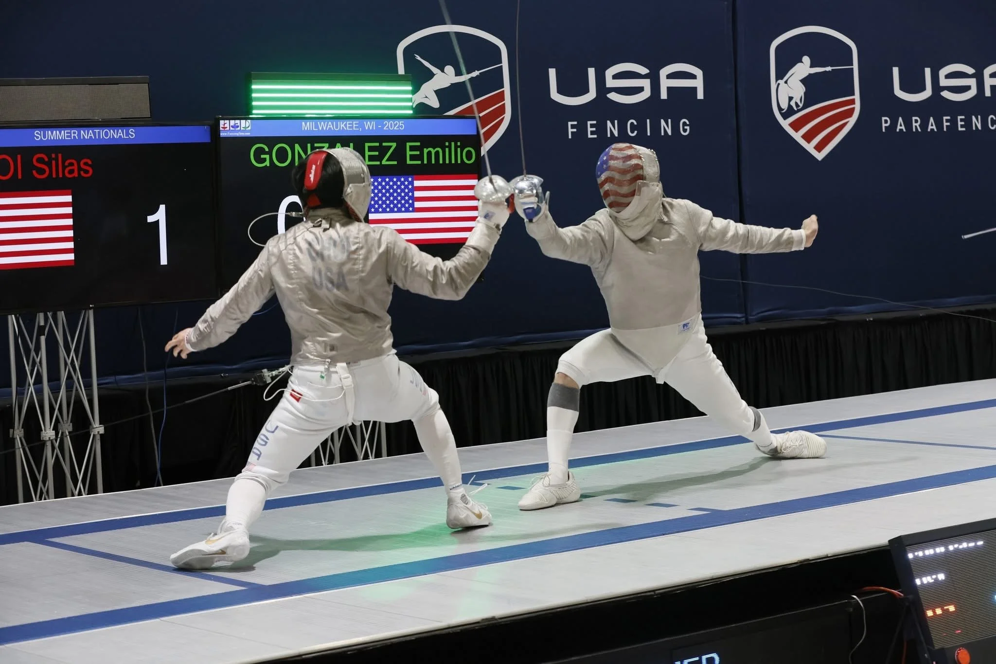 Two fencers in white gear sparring during a competition, with a digital scoreboard and USA fencing banners in the background.