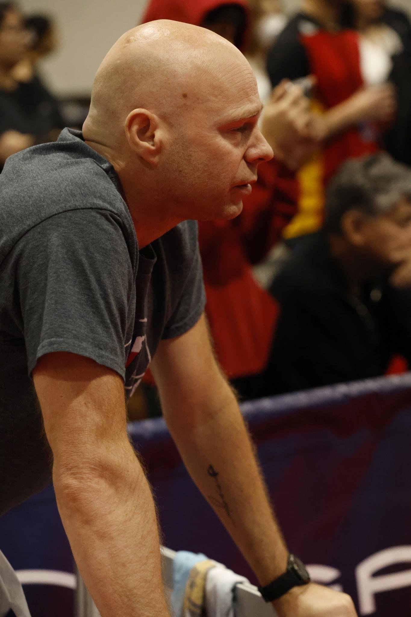 A bald man in a gray T-shirt leaning forward, surrounded by a crowd of people in a setting that appears to be a sports event or a public gathering.