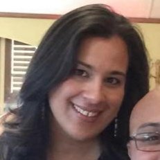 A woman with dark hair smiling, partially visible person next to her, indoor setting with a window in the background.