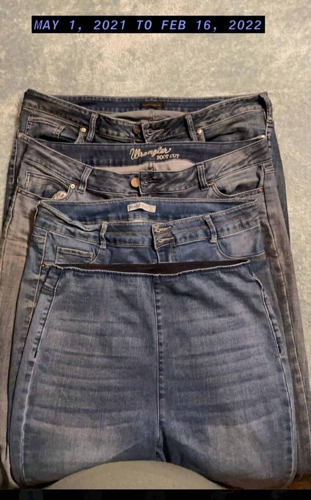 Five pairs of blue jeans laid flat on the floor, stacked on top of each other. The jeans vary in shades of blue and style, with some labeled 'Wrangler,' 'Hudd,' and others, showing different washes and fits. A date range "May 1, 2021 to Feb 16, 2022" is displayed at the top of the image.
