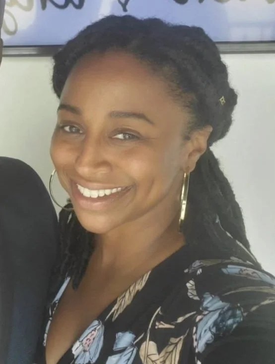 A smiling Black woman with dreadlocks, wearing large hoop earrings and a floral top.