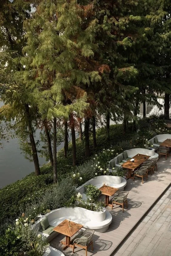 Waterside dining terrace with sculptural curved planters, teak tables, and woven chairs set among tall evergreens, boutique hospitality and landscape design.