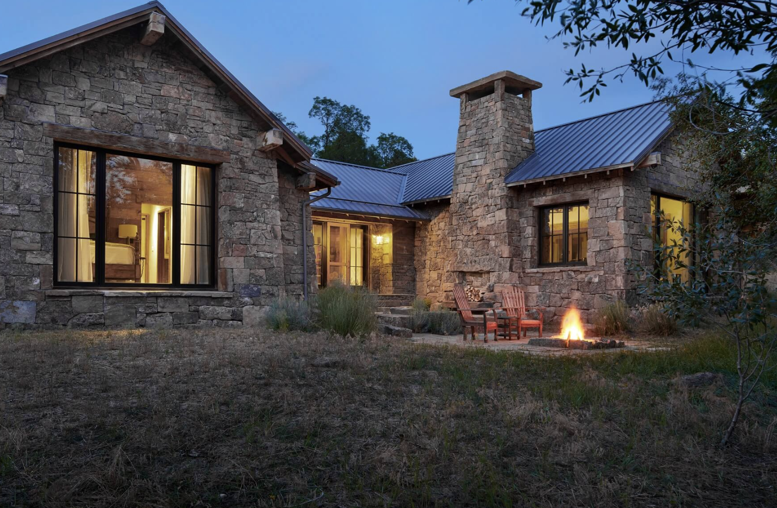 Stone mountain home at dusk with a metal roof, glowing windows, and an outdoor firepit, illustrating the relationship between architecture and interior design.