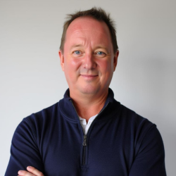 A man with short dark hair wearing a navy blue zip-up sweater, standing against a light gray background, smiling with arms crossed.