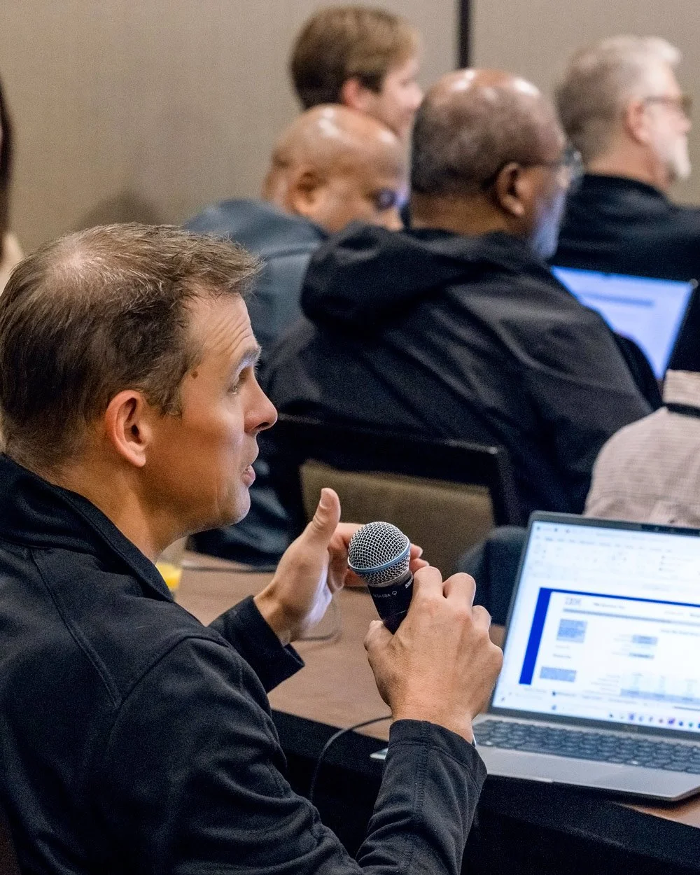 A man in a black jacket holding a microphone and speaking into it during a conference or meeting, with a laptop open in front of him and other people sitting nearby listening.