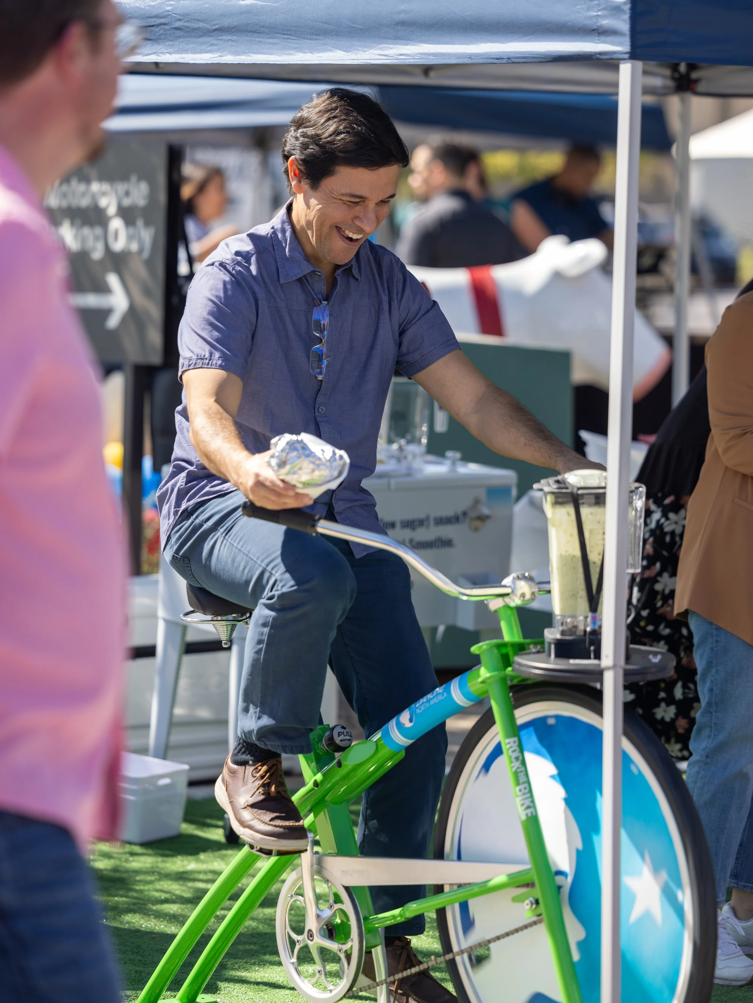 A man riding a green and blue stationary bicycle with a smile at an outdoor event or market.