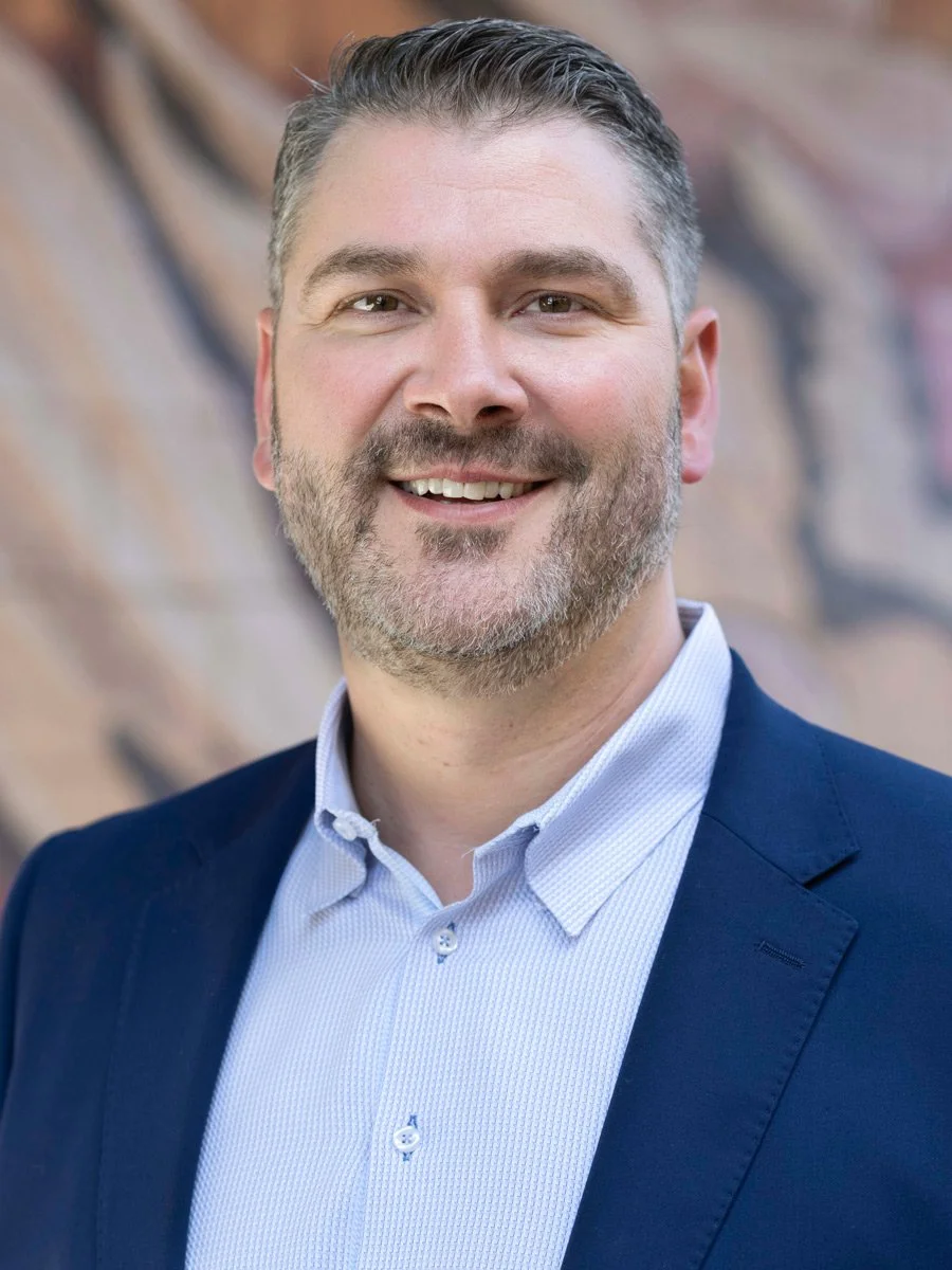 Portrait of a man with short, dark hair and a beard, smiling, wearing a light blue dress shirt and a navy blazer, standing outdoors with a blurred brick wall background.
