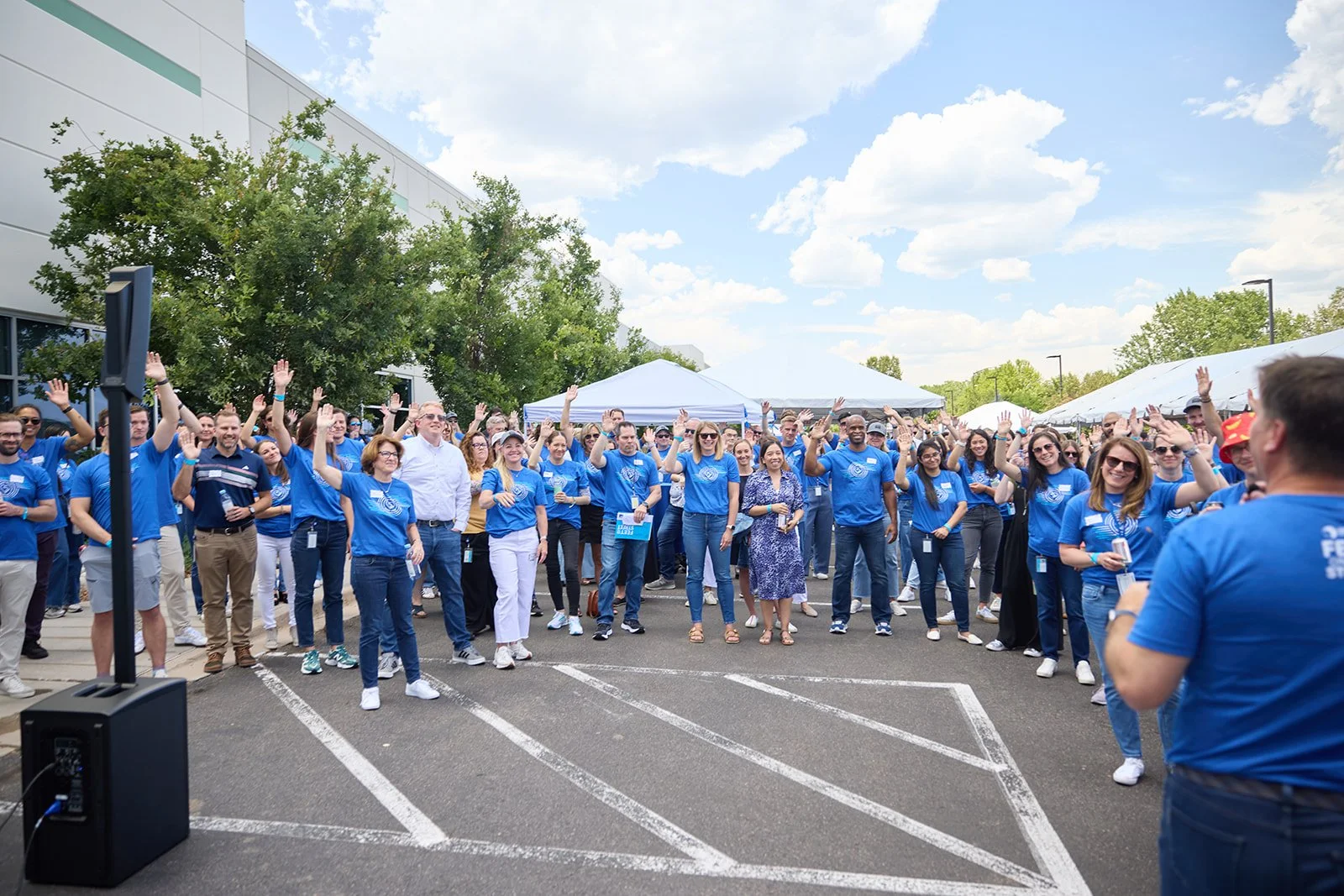 A large group of people outdoors, many wearing blue shirts with a logo, gathered under tents on a parking lot, waving and smiling during a daytime event on a partly cloudy day.