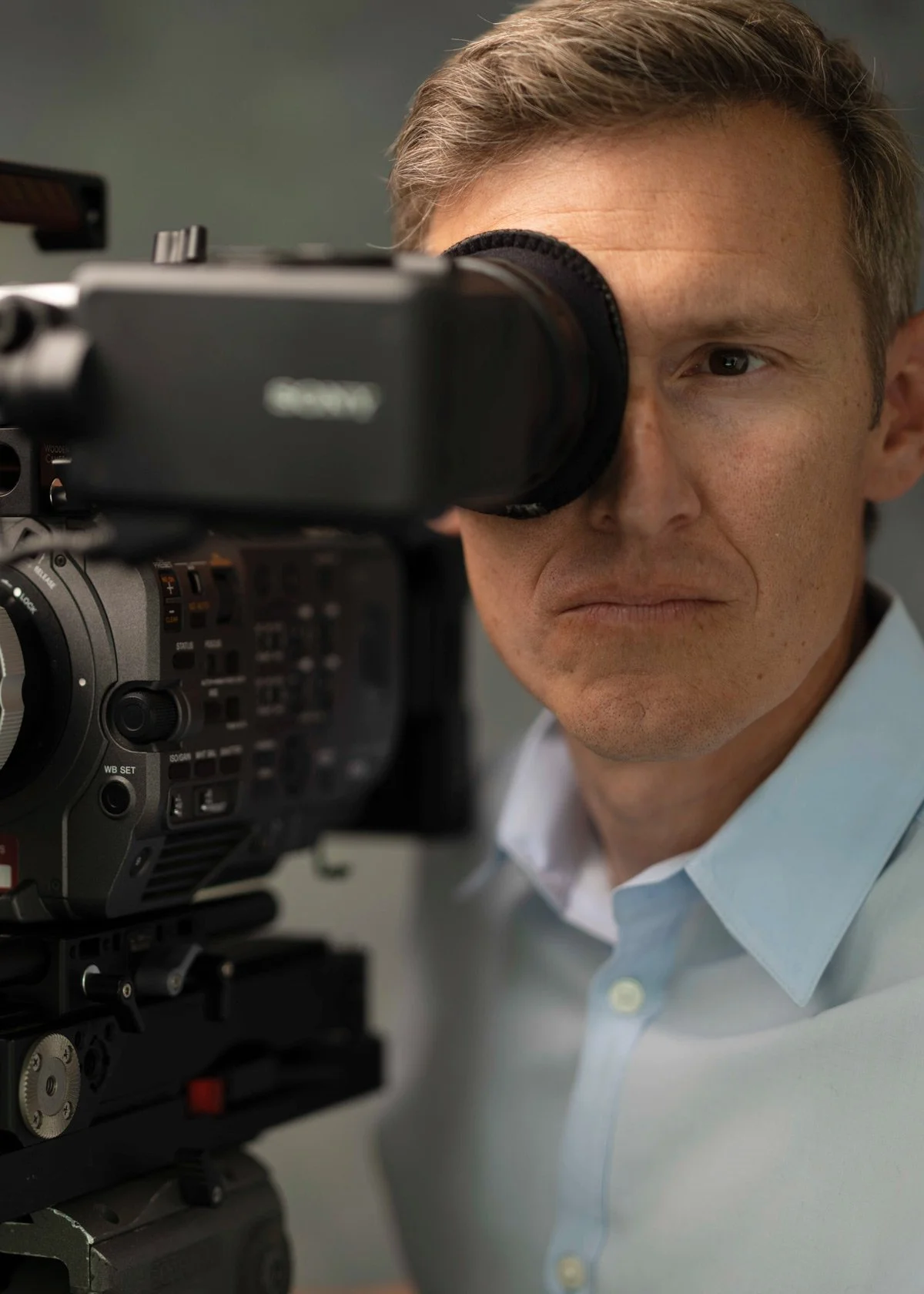A man in a light blue shirt looking through the viewfinder of a professional video camera.