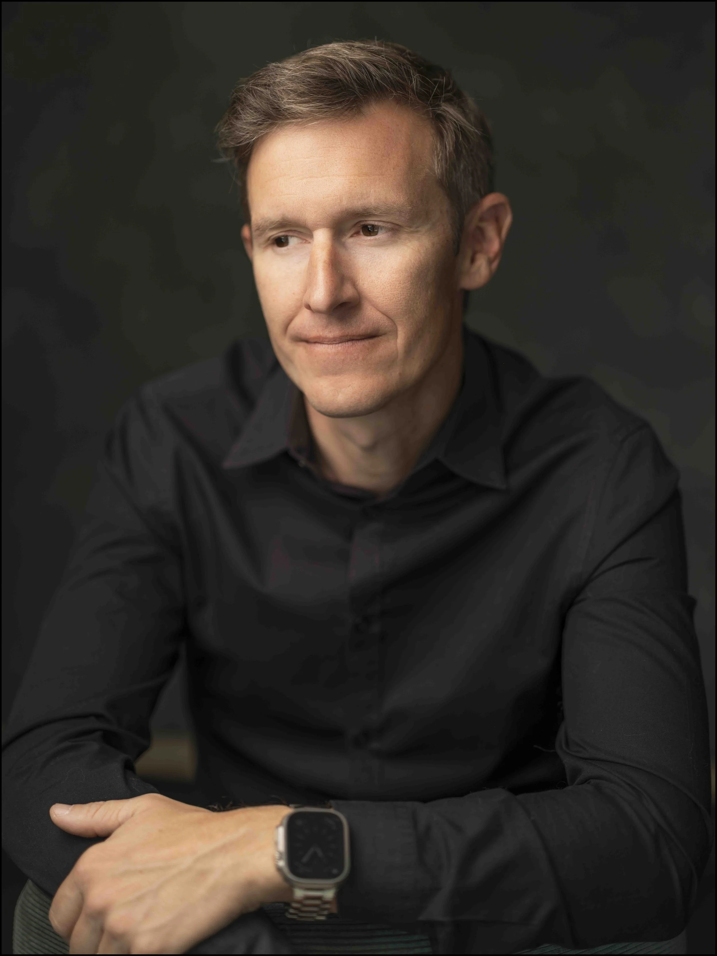 A man in a black shirt sitting with arms crossed, wearing a smartwatch, and looking thoughtfully to his right against a dark background.