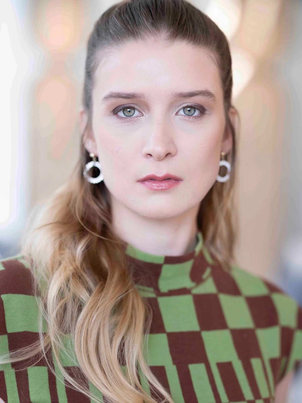 Portrait of a young woman with blue eyes, long wavy hair, wearing earrings and a green and brown checkered top.