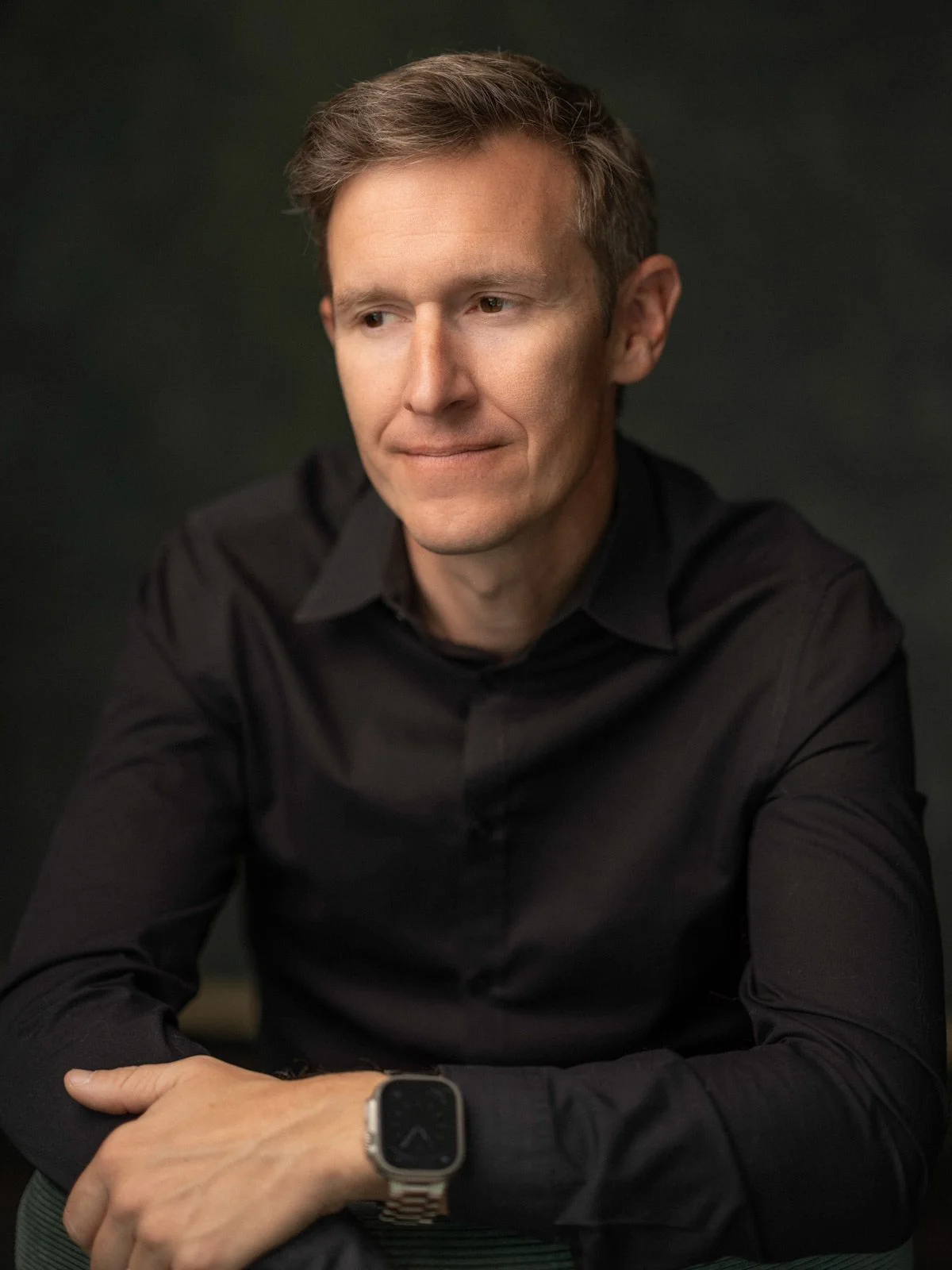 Portrait of a man in a black shirt with a smartwatch on his wrist, sitting with his arms crossed, looking to the side, against a dark background.