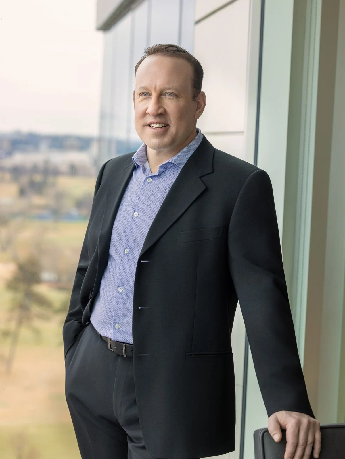 A man in formal business attire standing next to a window with a cityscape view, smiling and looking at the camera.