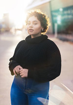 A woman standing outdoors in sunlight, wearing a black sweater and blue jeans, with her hands clasped in front of her.