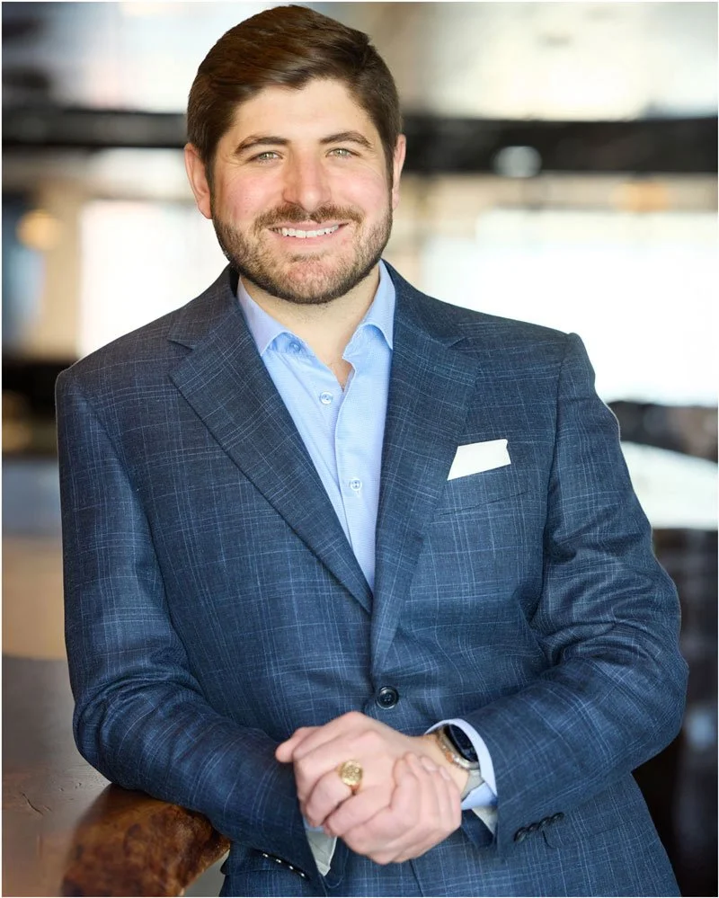 A man dressed in a blue checked suit with a light blue shirt, smiling with his hands clasped, in a professional setting.