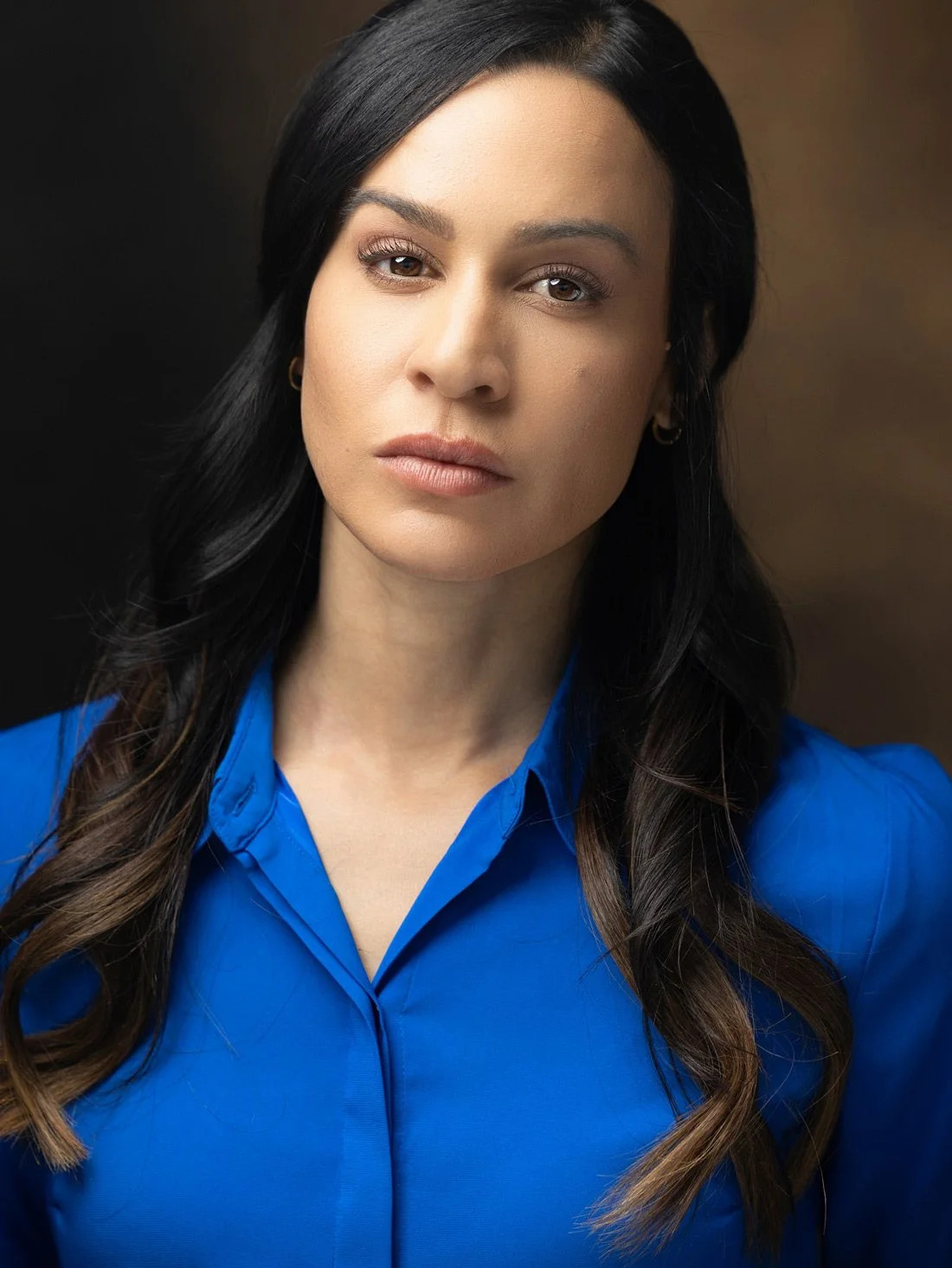 A woman with long dark hair wearing a bright blue blouse, looking directly at the camera with a neutral expression, against a dark background.