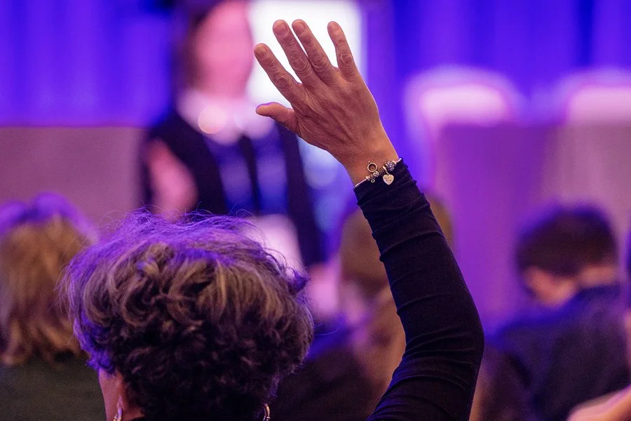 A person with short curly hair raising their hand in a crowded indoor setting with purple lighting.