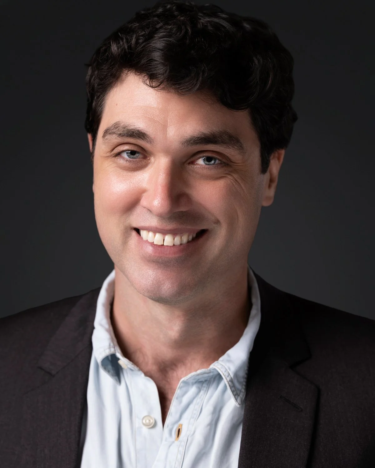 Headshot of a man with dark curly hair, blue eyes, wearing a dark blazer and white shirt, smiling against a dark background.