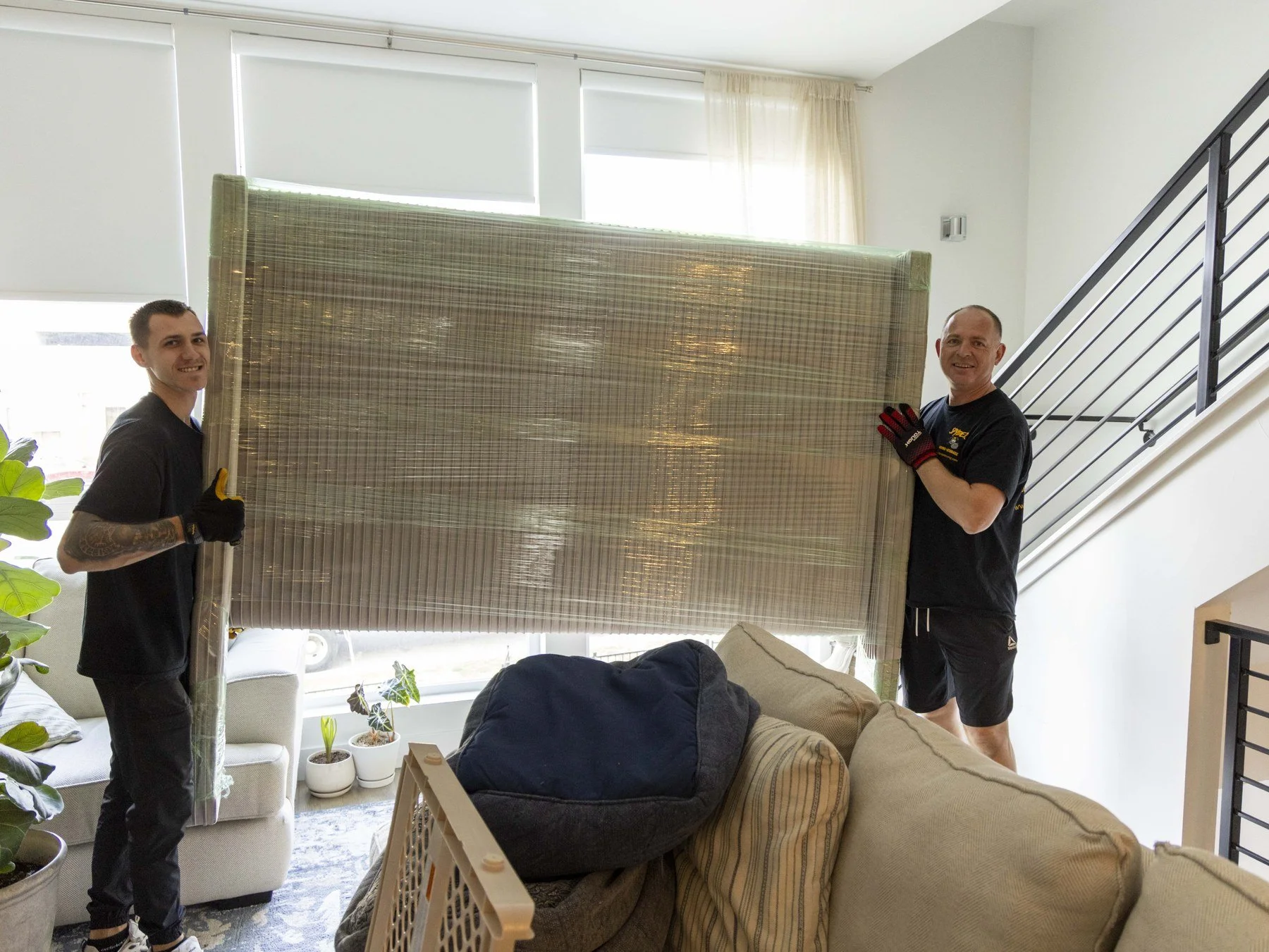 Two men hold a large, wrapped object inside a bright living room. One man is on the left with tattoos on his arms, and the other on the right wears gloves. The room has windows with blinds, potted plants, a sofa, and a staircase.