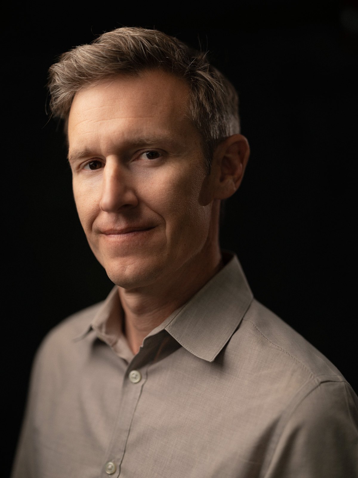 Portrait of a man with light brown hair, wearing a beige dress shirt, against a dark background.