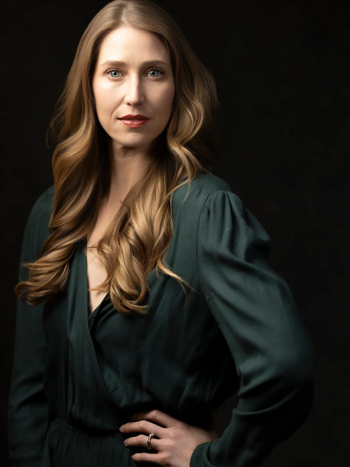 A woman with long, wavy, blonde hair and blue eyes, wearing a dark teal jacket and gold ring, standing against a dark background.