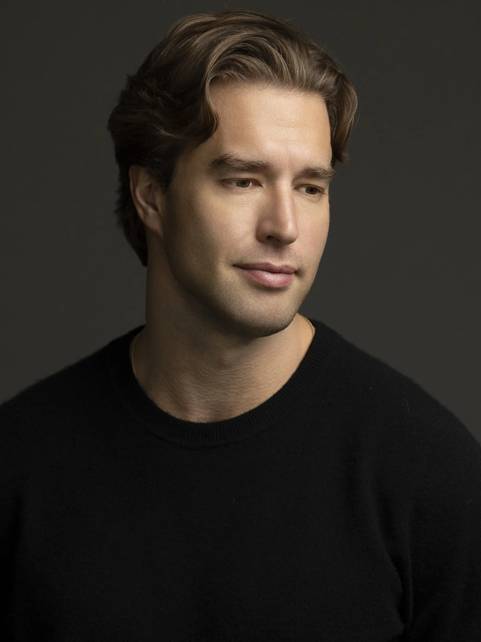 A young man with brown hair and fair skin, wearing a black sweater, looking down with a slight smile against a dark background.