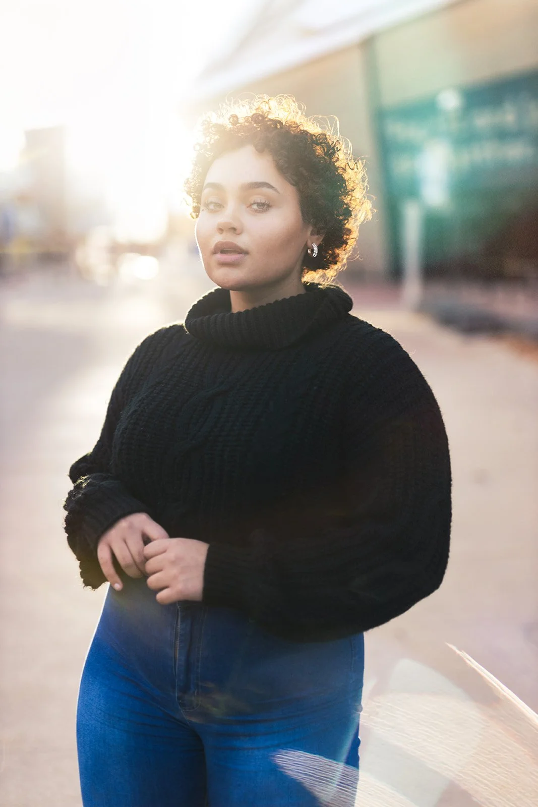 Young woman with short, curly hair standing outdoors at sunset, wearing a black turtleneck sweater and blue jeans.